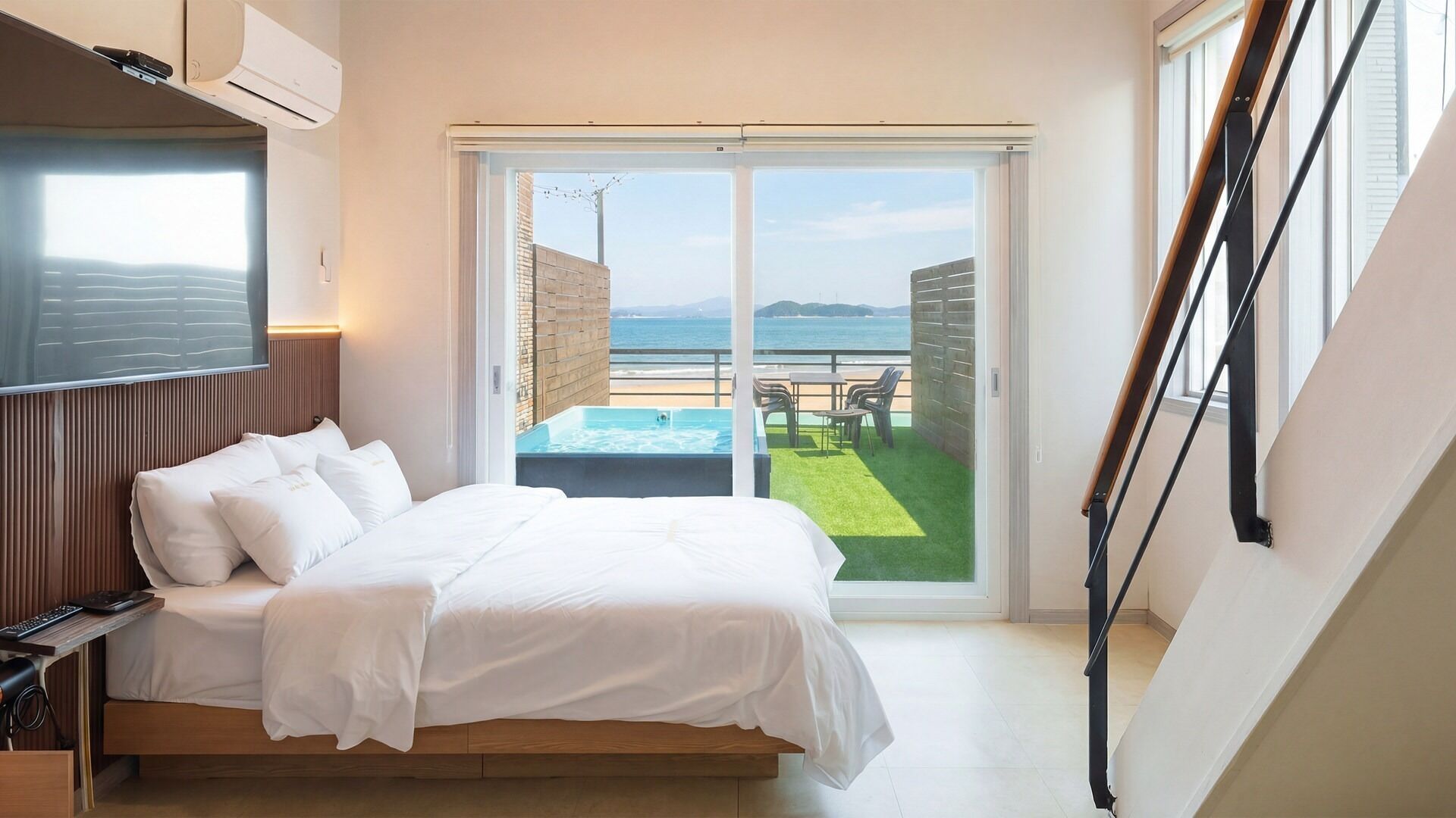 Un dormitorio con cama, televisión y puerta corrediza de vidrio que da a un balcón con piscina y vista al mar.