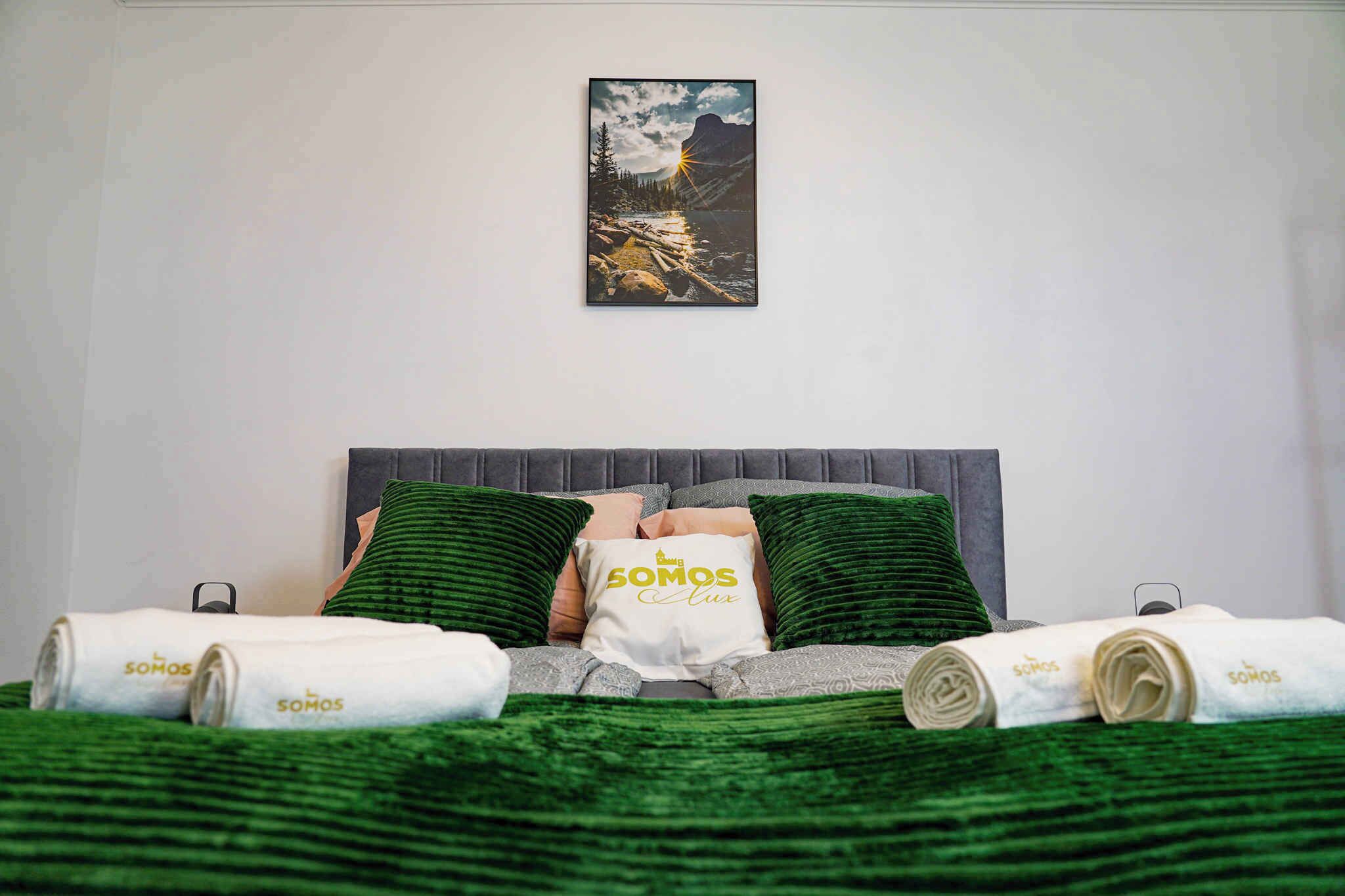 Una cama bien hecha con ropa de cama verde y blanca, un cojín con el texto "Somos Lux" y un cuadro de un paisaje montañoso encima.