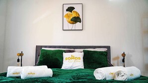 Een net opgemaakt bed met groen beddengoed, een kussen met de tekst "SOMOS Lux" en twee opgerolde handdoeken.