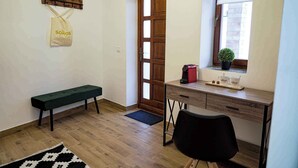 Een kamer met een houten bank, een bureau met een computer, een kleine plant in pot en een raam met jaloezieën.