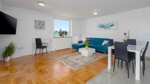 Uma sala de estar moderna com um sofá azul, mesa de centro branca e uma área de jantar com mesa branca e cadeiras pretas.