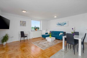 A modern living room with a blue sofa, white coffee table, and a dining area with a white table and black chairs. - Holiday apartment with air conditioning and balcony (Funtana)