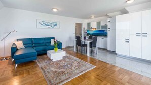 Uma sala de estar moderna com um sofá azul, uma mesa de centro branca e um tapete estampado. Há um espaço de jantar com mesa e cadeiras, e uma cozinha com armários brancos e um revestimento azul.
