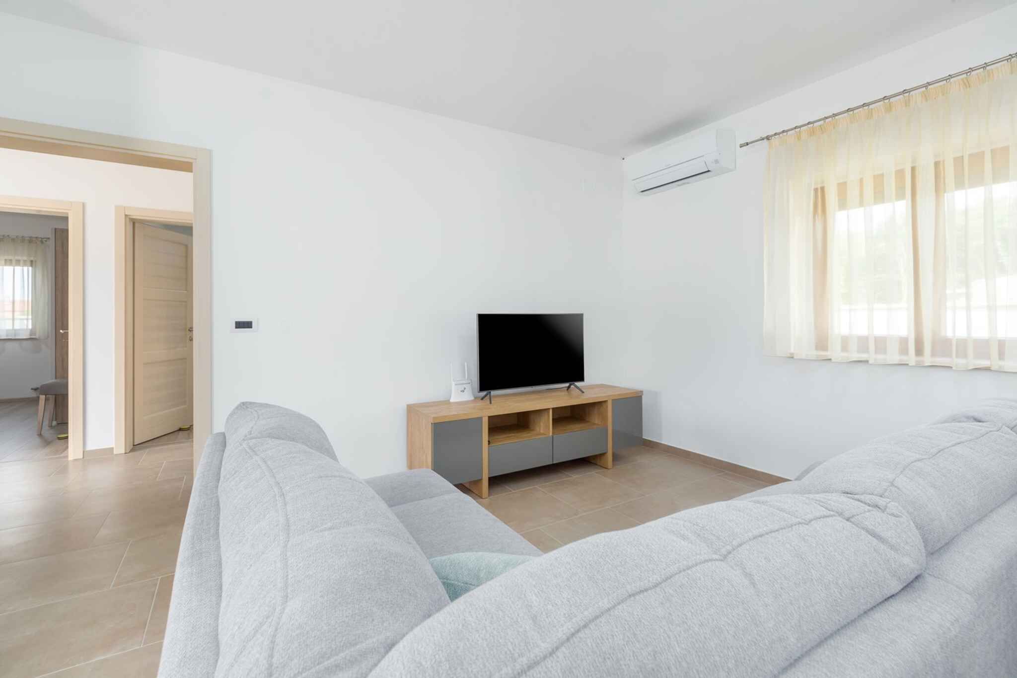 A modern living room with a grey sofa, a flat-screen TV on a wooden stand, and a window with sheer curtains.