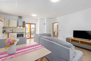 A modern living room with a grey sofa, a dining table with a striped table runner, and a flat-screen TV on a wooden stand. - Villa with air conditioning and pool (Kringa)