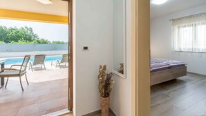 Una habitación con cama, ventana con cortinas, un espejo pequeño montado en la pared, y vistas a una piscina y sillas afuera.