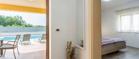 A room with a bed, a window with curtains, a small wall-mounted mirror, and a view of a swimming pool and chairs outside.