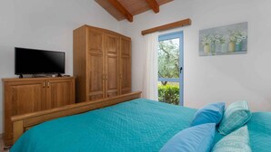 Ein Schlafzimmer mit einem großen Bett, einem Holzschrank, einem Fernseher auf einem Standfuß und einem Fenster mit Blick auf Grünflächen.
