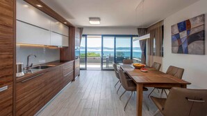 A modern kitchen with a large island, a dining area with a wooden table and chairs, and a view of the sea through large windows. - Holiday apartment with shared-use pool (Sv. Filip i Jakov)