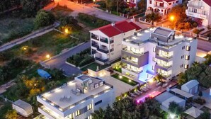 A modern multi-story building with a flat roof, large windows, and a well-lit exterior. - Holiday apartment with shared-use pool (Sv. Filip i Jakov)