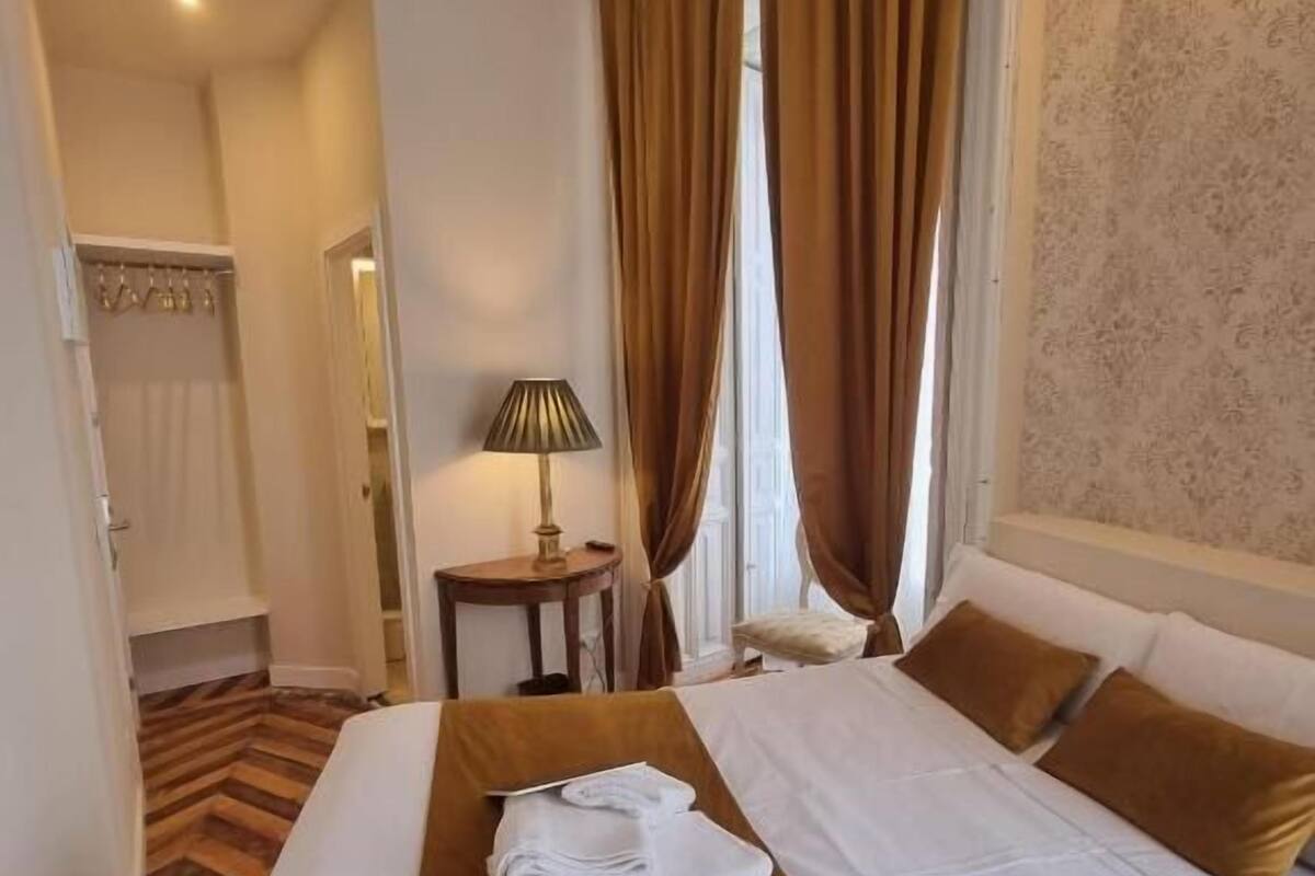 A hotel room with a bed, a bedside table with a lamp, a window with curtains, and a wooden floor.