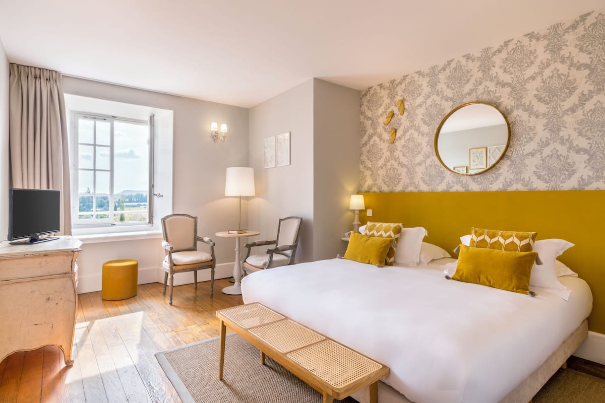 A hotel room with a large bed, a desk with a television, a chair, a small table, and a window with curtains.