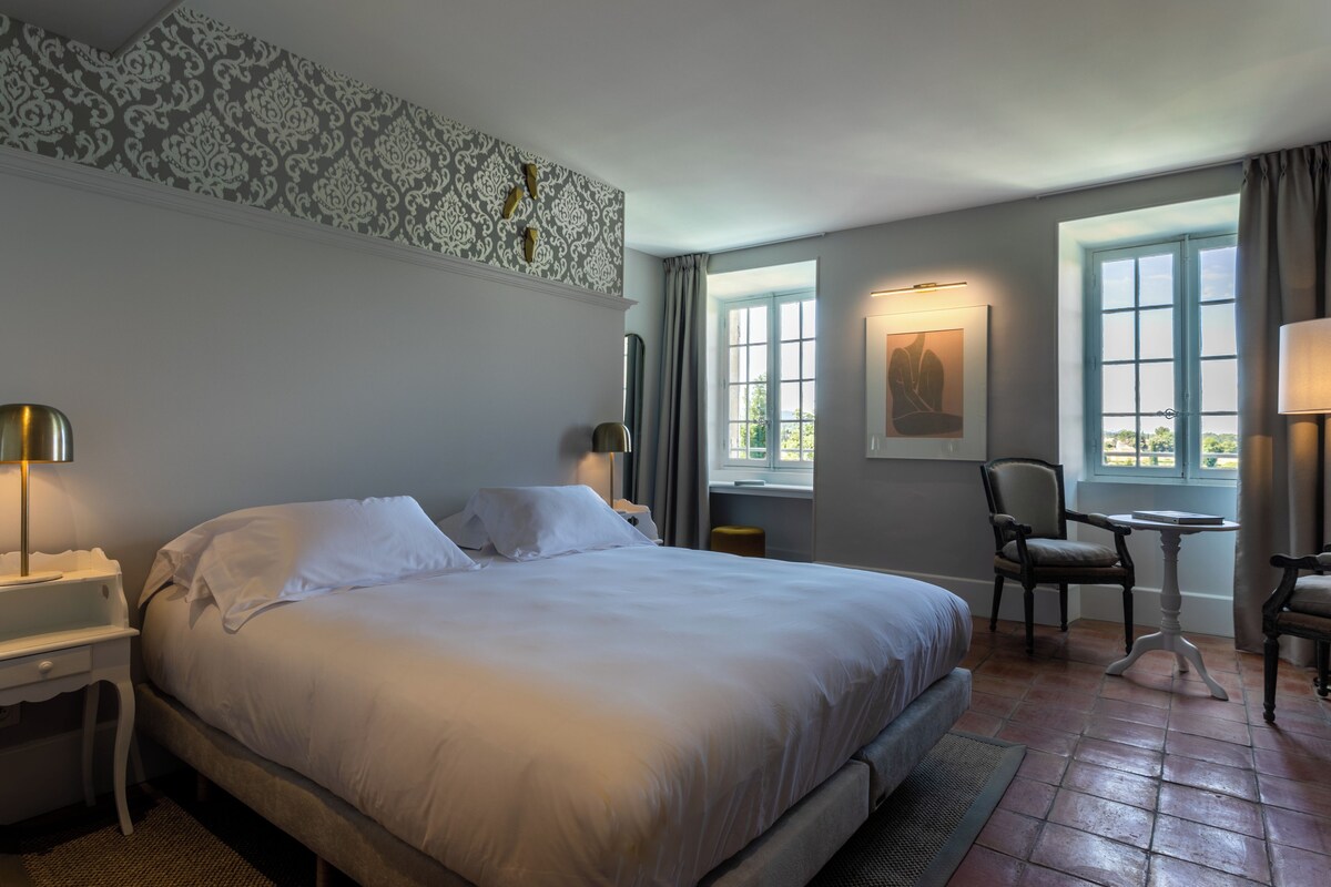 A hotel room with a large bed, two bedside tables, a chair, a small table, and a window with curtains.