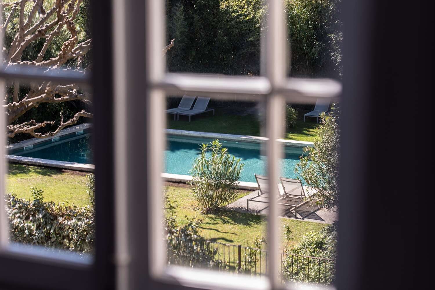 Vista a través de una ventana de una piscina con sillones de descanso, rodeada de vegetación.