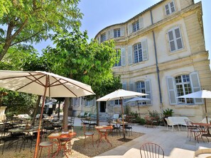 Un espace de café en plein air avec des tables et des chaises, des parasols, et un bâtiment avec de nombreuses fenêtres et des volets en arrière-plan.