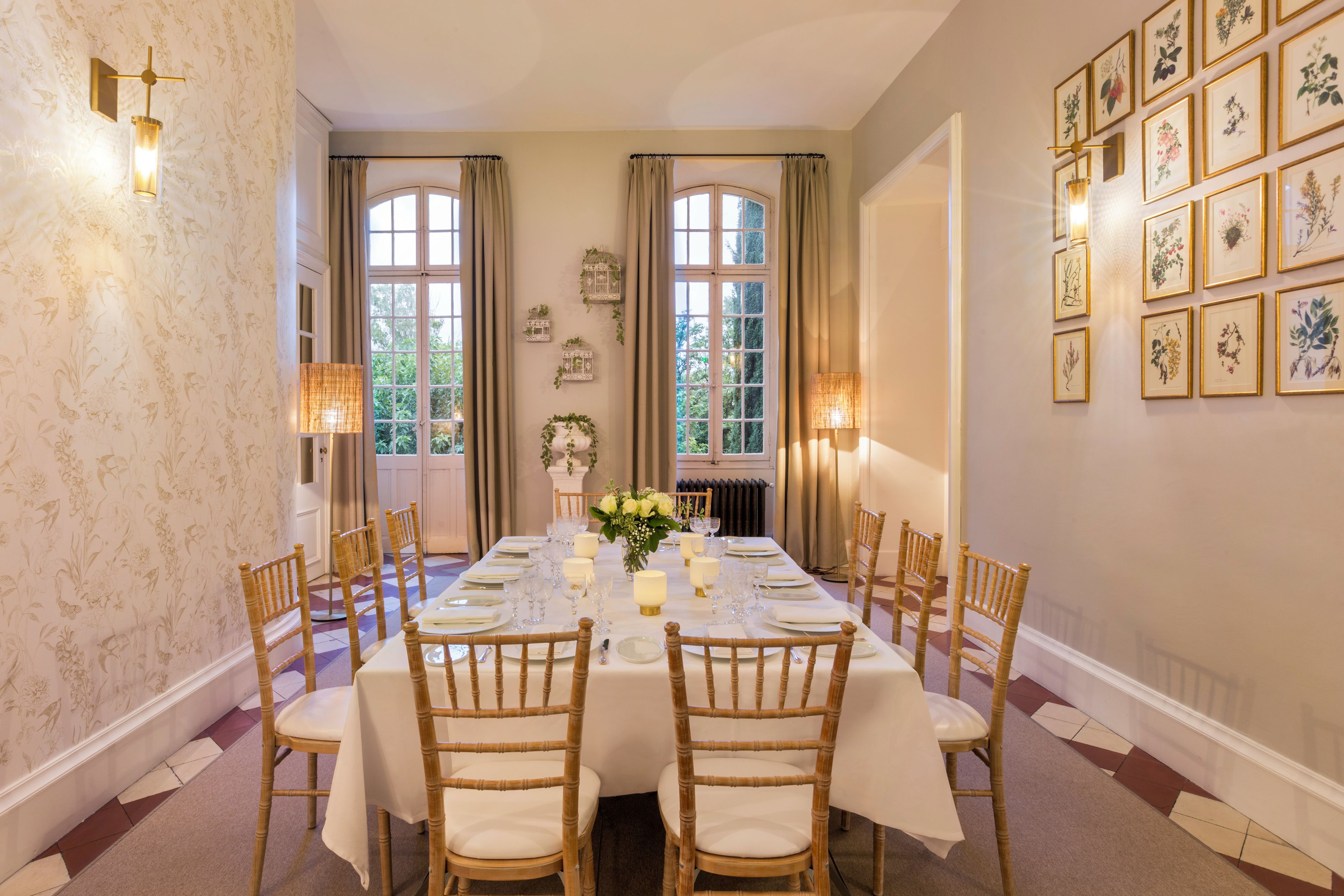 Comedor formal con una larga mesa dispuesta para seis comensales, flanqueada por altas ventanas con cortinas, y adornado con grabados botánicos en las paredes.