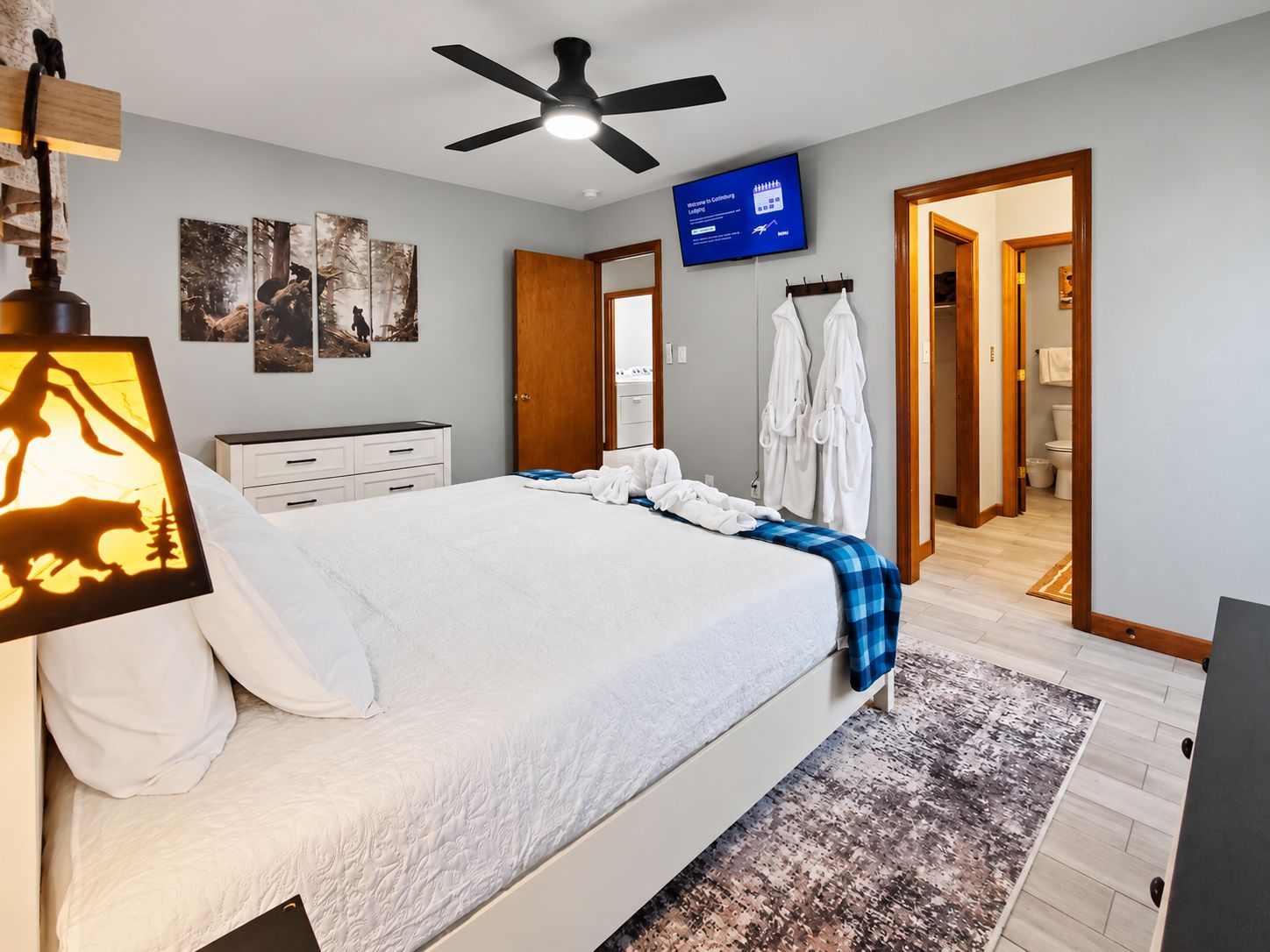 A hotel room with a bed, a ceiling fan, a television, artwork on the wall, and a bathroom visible through an open door.