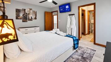 A hotel room with a bed, a ceiling fan, a television, artwork on the wall, and a bathroom visible through an open door.