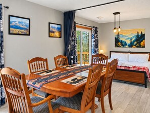 Un coin repas avec une table et des chaises en bois, une vue sur les montagnes par la fenêtre, et un lit avec une tête de lit dans la pièce adjacente.