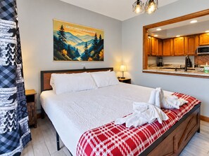 Une chambre d’hôtel avec un grand lit, un tableau représentant les montagnes Great Smoky, une tête de lit en bois, une table de chevet avec une lampe et un coin cuisine visible à travers un miroir.