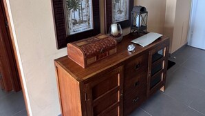 Un mueble de madera con cajones y una mesita, un cuadro enmarcado con palmeras en la pared, y una puerta al fondo.