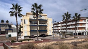 Un muelle de madera que conduce a una zona de playa con palmeras y edificios al fondo.