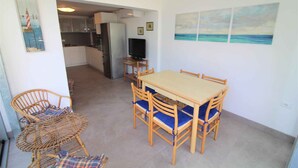 A dining area with a wooden table and chairs, a television on a stand, and a kitchen in the background. - Holiday apartment with common garden (Lignano Sabbiadoro)