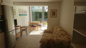 A compact living space with a kitchenette, a sofa, a dining table with chairs, and a large sliding glass door leading to an outdoor area. - Holiday apartment with common garden (Lignano Sabbiadoro)