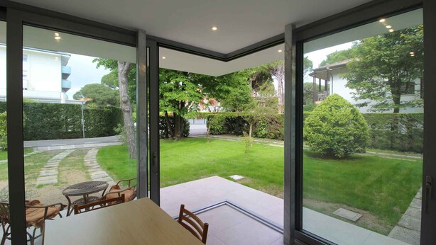 A modern room with a large sliding glass door leading to a well-maintained garden. - Holiday apartment with common garden (Lignano Sabbiadoro)