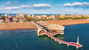 Un hotel sul lungomare con un lungo molo che si protende nell'acqua, una spiaggia con file di ombrelloni e uno skyline sullo sfondo.