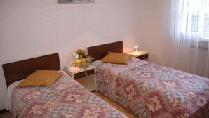 Una stanza con due letti, ciascuno con una testiera in legno e una coperta a fiori. Si nota un comodino con una lampada e un tavolino con un vaso di fiori.