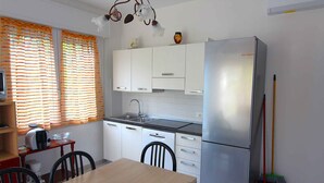 Una cocina compacta con armarios blancos, un refrigerador de acero inoxidable, una mesa de comedor con sillas y una ventana con cortinas a rayas.