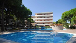 A swimming pool with a tiled deck, surrounded by trees and a multi-story building in the background. - Holiday apartment mit Pool (Lignano Sabbiadoro)