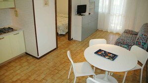 A compact kitchen with a gas stove, a dining table with chairs, a sofa, and a television on a cabinet. - Holiday apartment mit Pool und Balkon (Lignano Sabbiadoro)