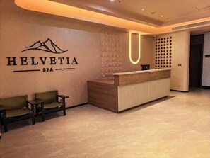 Un espace d’accueil avec un comptoir en bois, deux chaises vertes et un logo de « Helvetia Spa » fixé au mur.