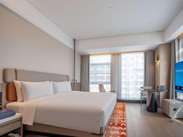 A hotel room with a bed, bedside table, chair, TV, and a window with curtains.
