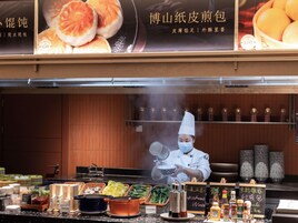 Montaje de bufé con diversos alimentos y condimentos, un chef con uniforme y un tablero con el menú.