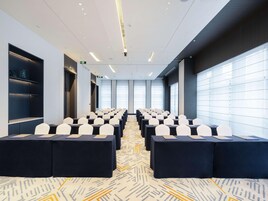 Un salón de conferencias con filas de mesas y sillas, ventanas amplias y una alfombra estampada.