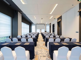 Un salón de conferencias con filas de mesas y sillas, ventanas amplias y una pantalla para proyector.