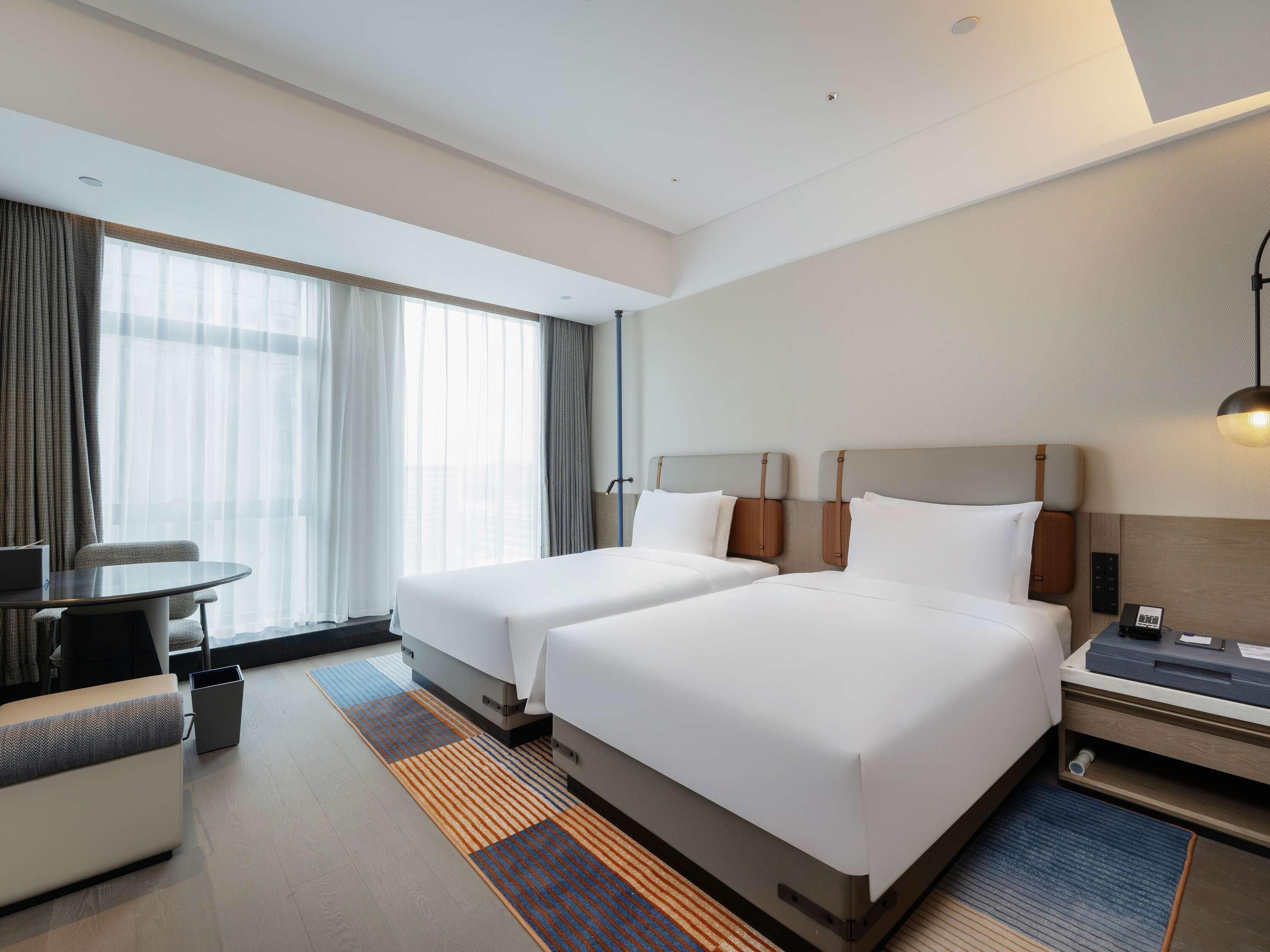 A hotel room with two beds, a desk, a chair, a lamp, and a large window with curtains.