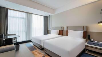 A hotel room with two beds, a desk, a chair, a lamp, and a large window with curtains.