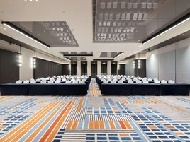 Un salón de conferencias con filas de mesas y sillas, una alfombra estampada y un diseño de techo moderno.