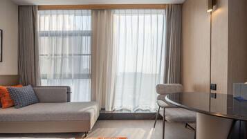 A modern hotel room with a sofa, a chair, a glass table, and a large window with curtains.