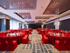 Un salón de conferencias con sillas y mesas rojas, una alfombra estampada y una pantalla grande al frente.