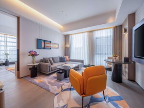 A modern hotel room with a sofa, coffee table, orange armchair, TV, and artwork on the wall.