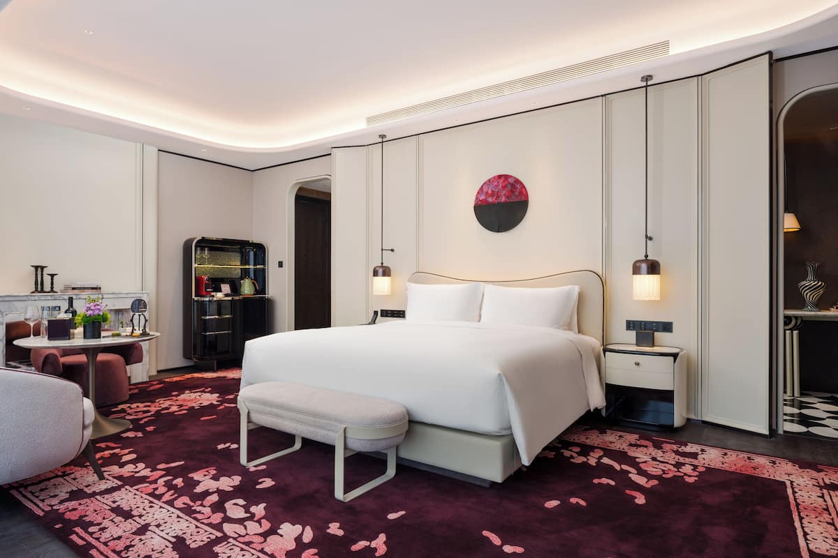 A modern hotel room with a large bed, bedside tables, a desk with a chair, a round mirror, and a decorative wall piece.
