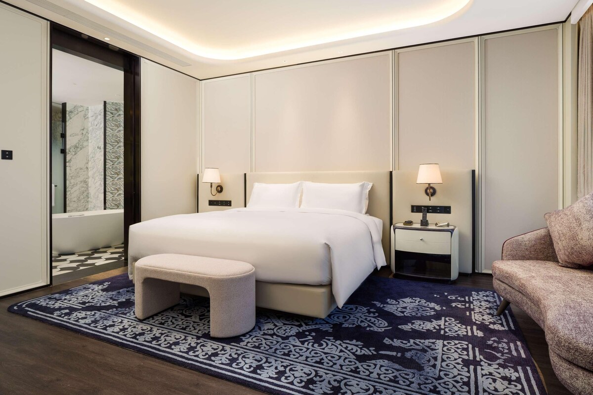 A modern hotel room with a large bed, bedside tables with lamps, a small stool, and a patterned rug.