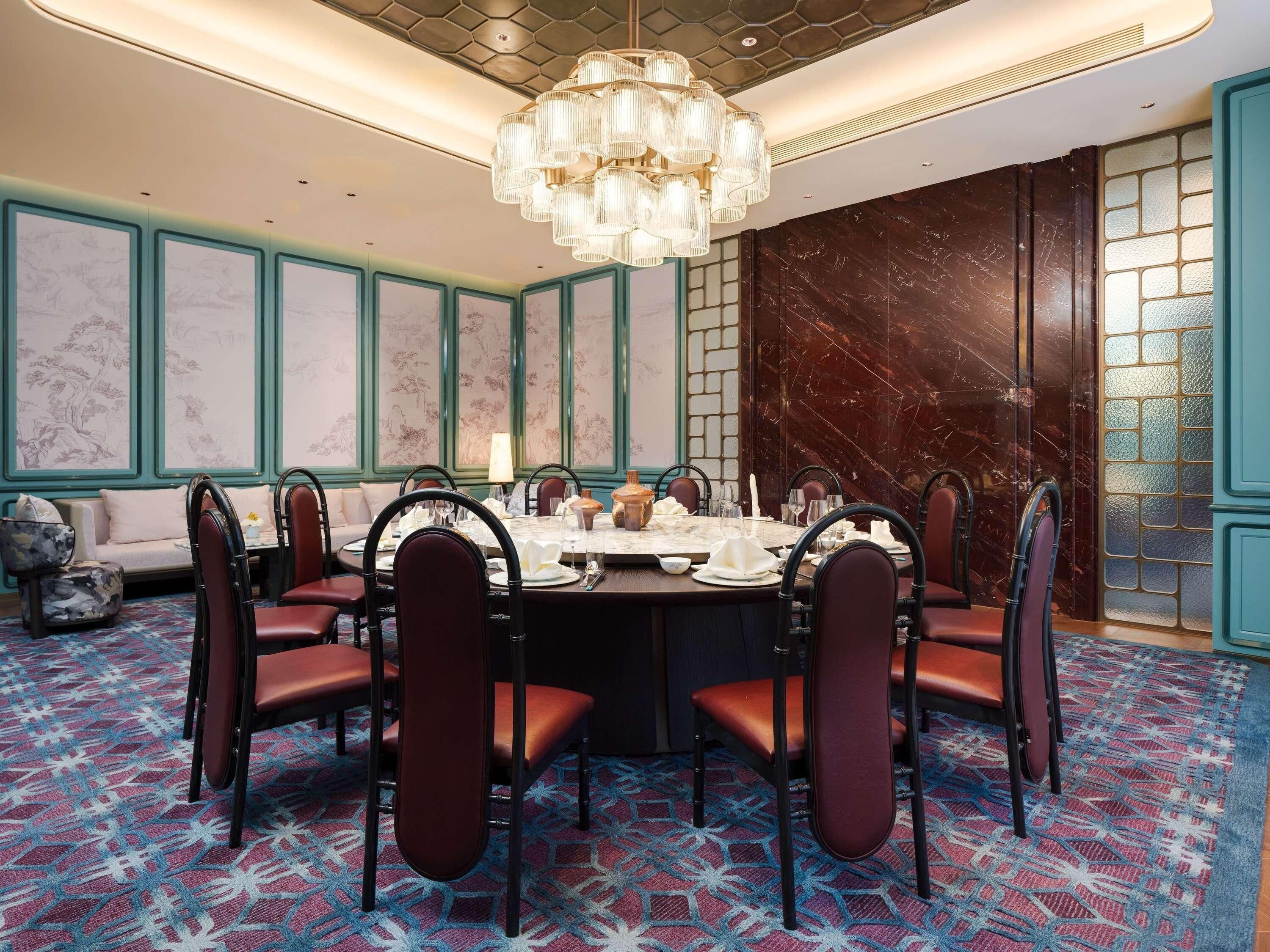Comedor con una mesa redonda dispuesta para seis personas, rodeada de sillas tapizadas, en una habitación con alfombra estampada y paneles decorativos.