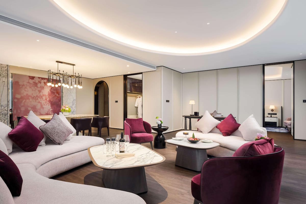 A modern hotel lobby with a curved ceiling, a seating area with sofas and armchairs, a dining table, and a large abstract painting.