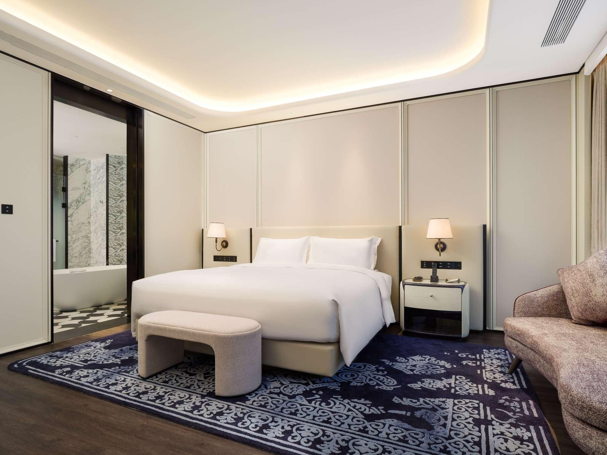 Una habitación de hotel moderna con una cama grande, mesitas de noche, un banco y una alfombra estampada.